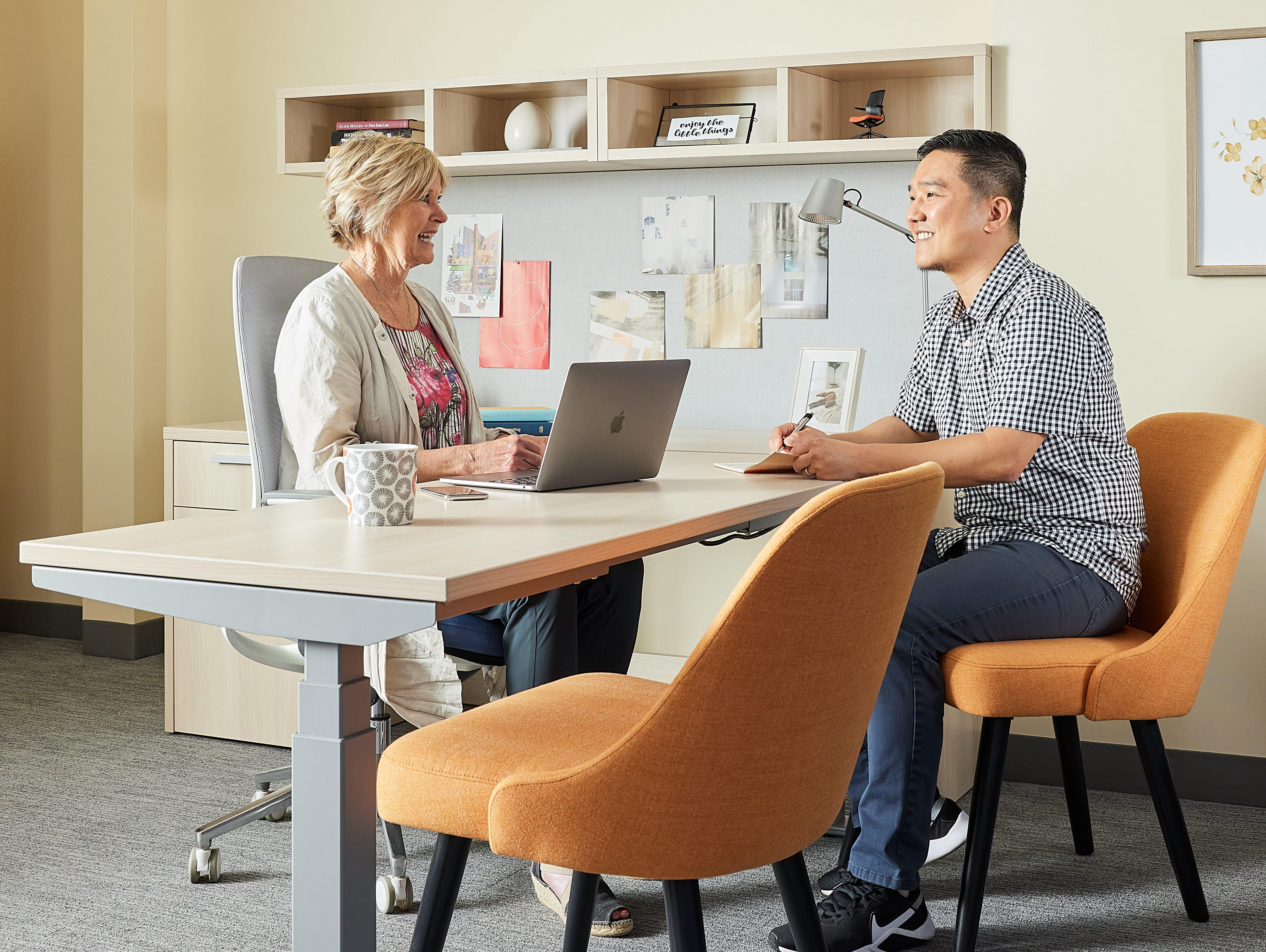Two colleagues chat at a height-adjustable office desk with laptop and notebook; orange upholstered chairs in a modern showroom office.