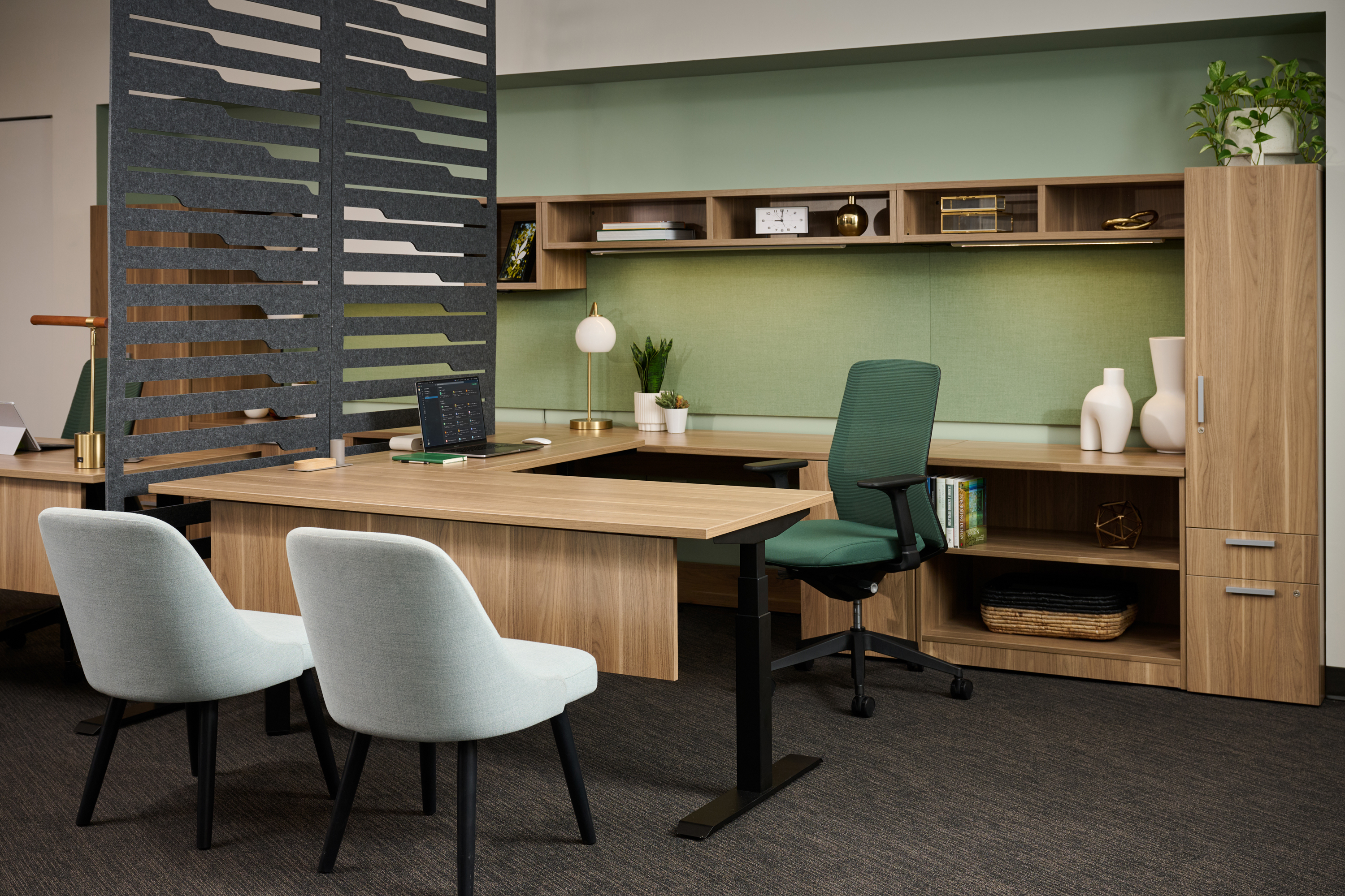 AMQ Solutions showroom: unoccupied wooden L-shaped desk with green ergonomic chair, two gray guest chairs, slatted partition, and wall shelving.
