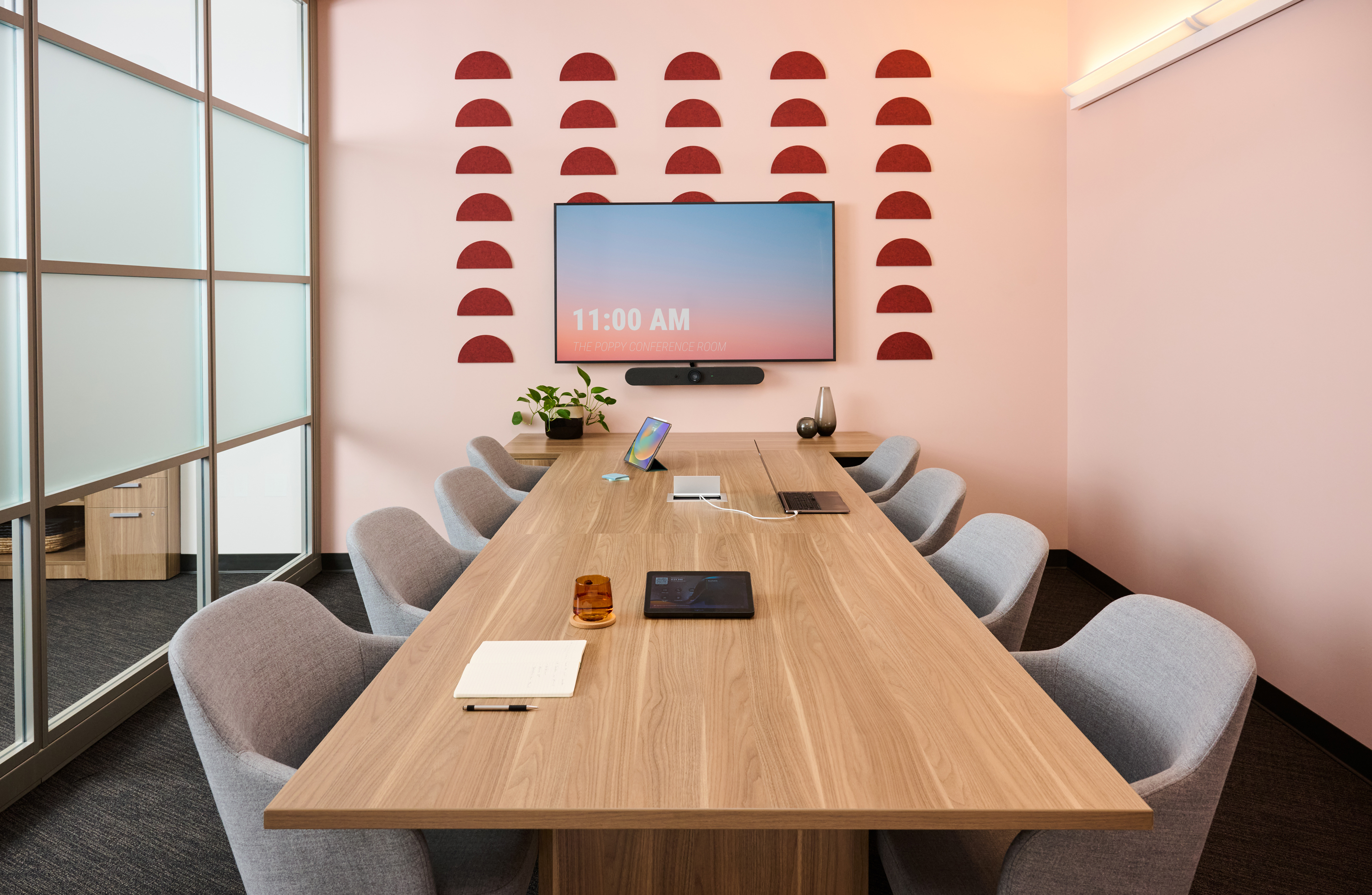 Long wooden conference table with gray upholstered chairs in a pink-walled meeting room, red semicircle wall art, and a wall-mounted screen.