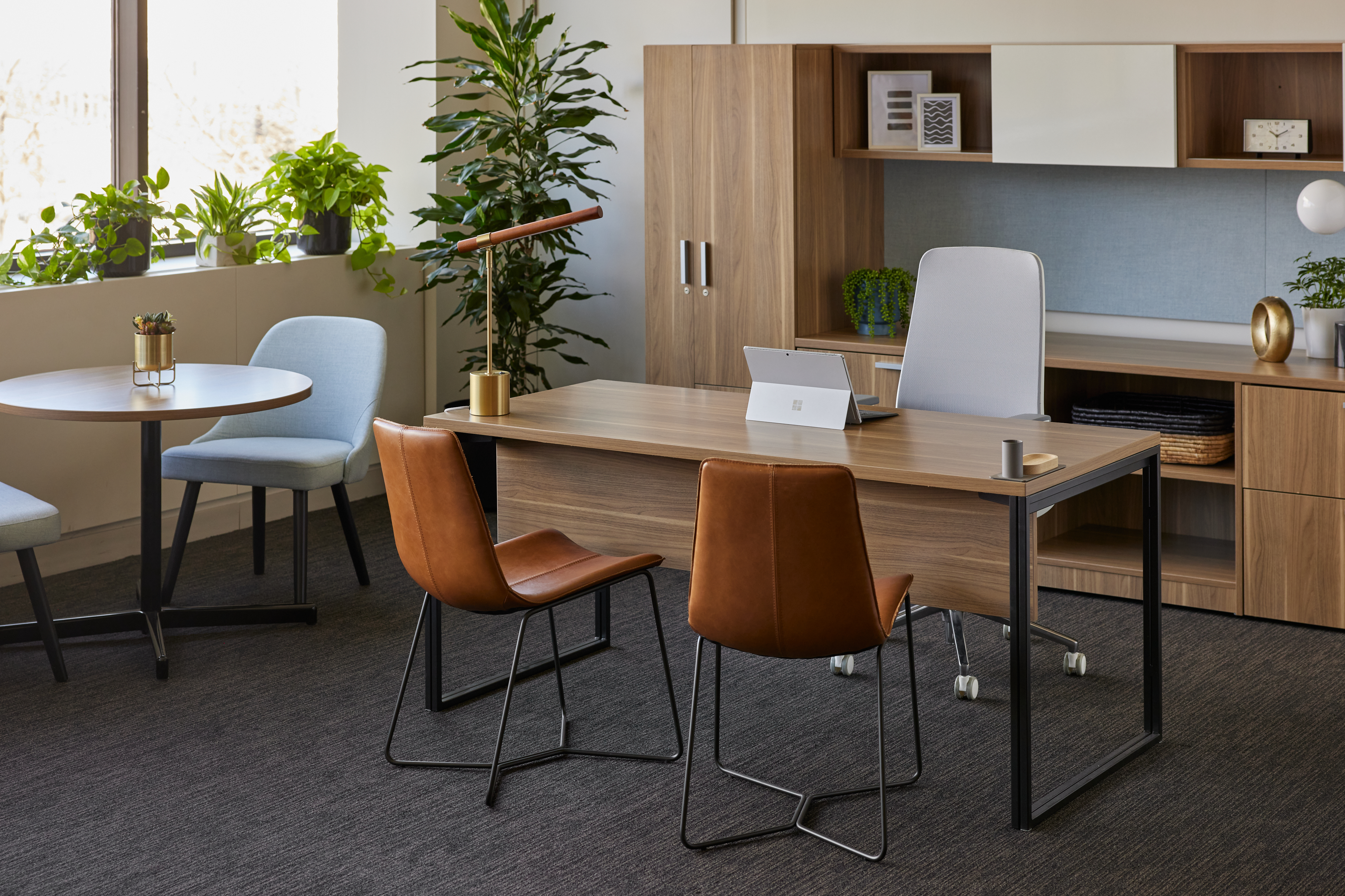 Wooden executive desk with integrated storage, white mesh chair, two brown leather guest chairs; potted plants and shelving.