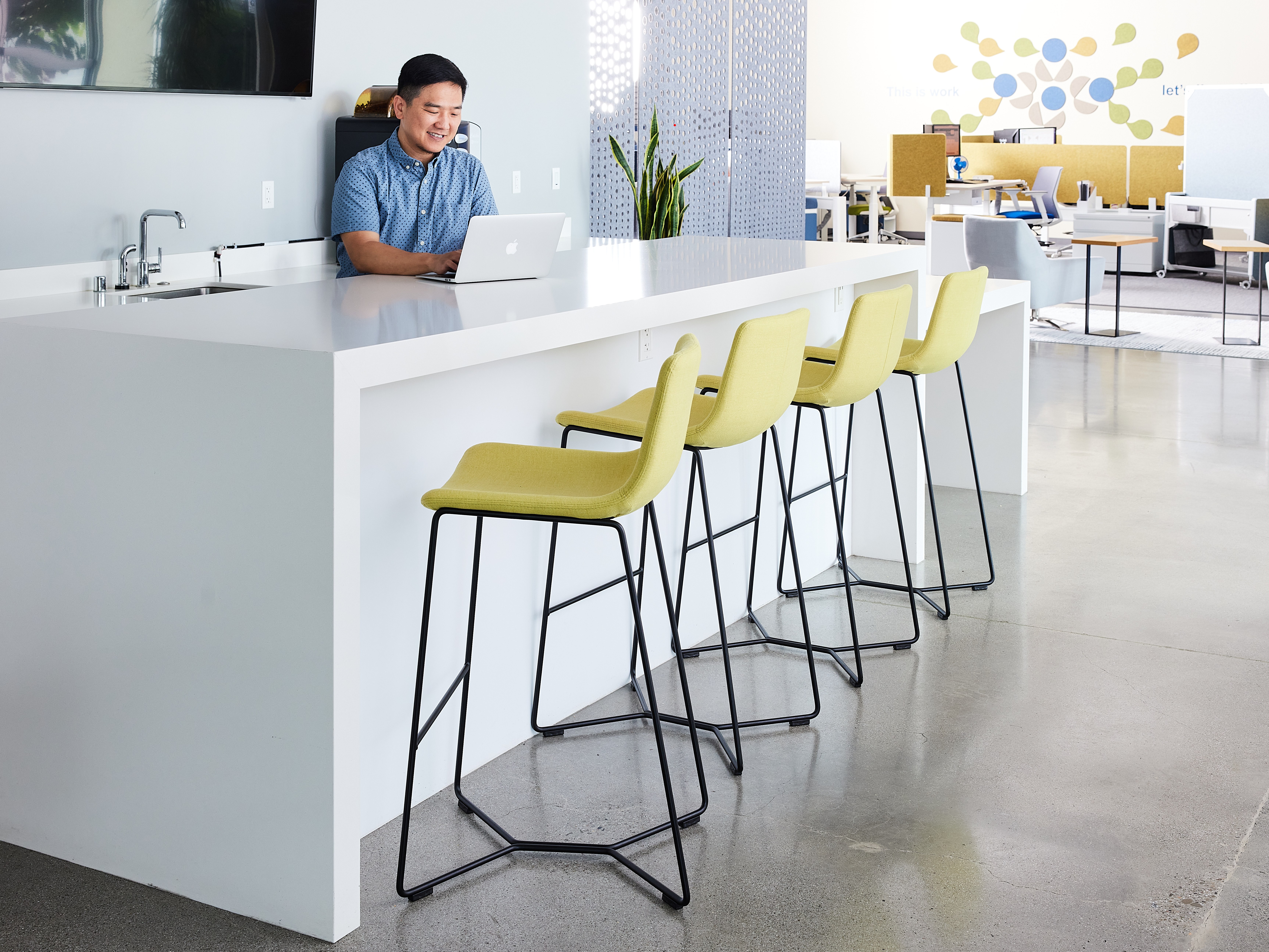 Person seated at white bar-height counter with laptop; yellow upholstered bar stools with black metal frames; open-plan office with plant and artwork.
