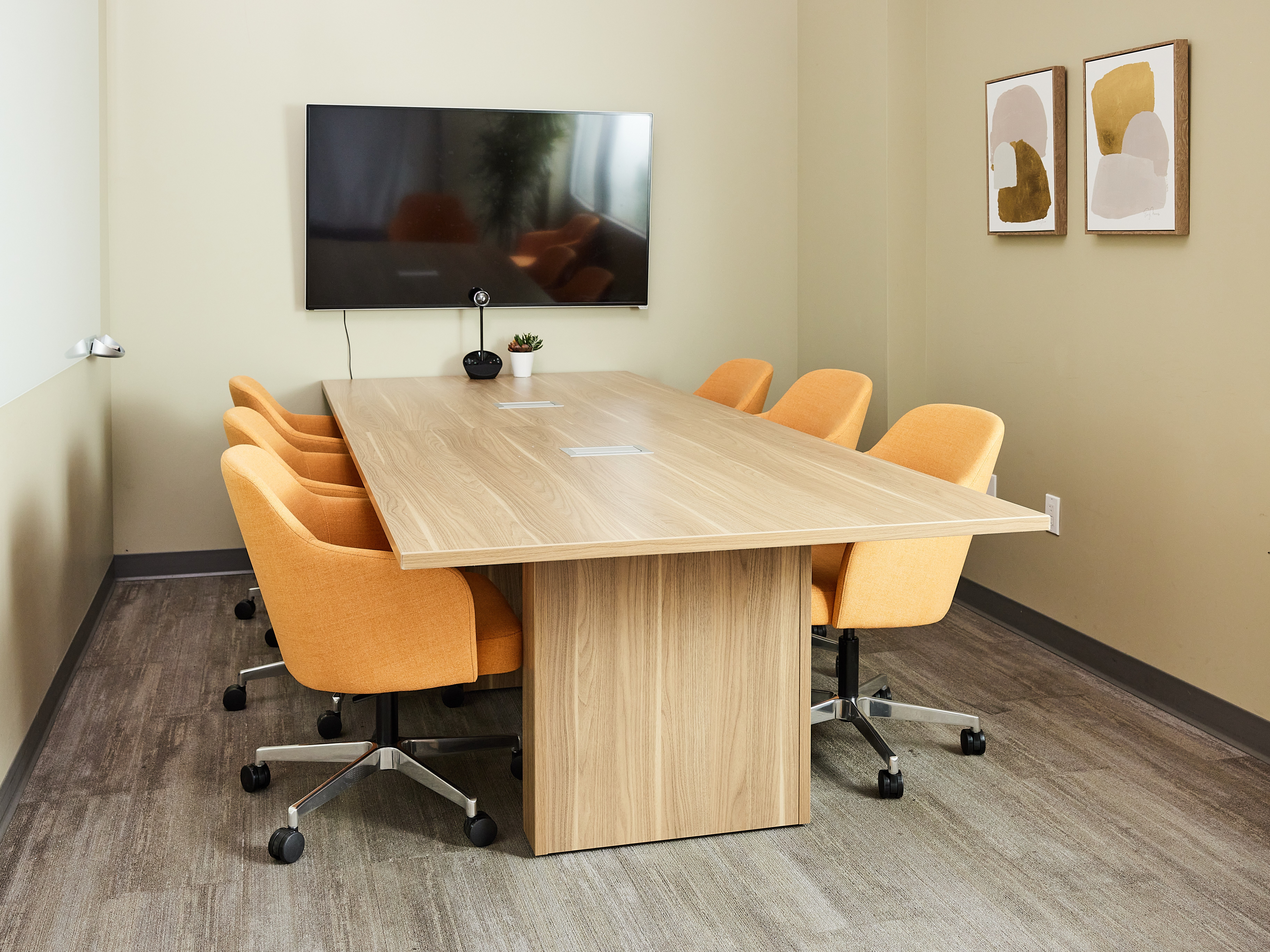 Long wooden rectangular conference table with orange swiveling chairs in a beige meeting room; wall-mounted screen, small plant, and abstract art.