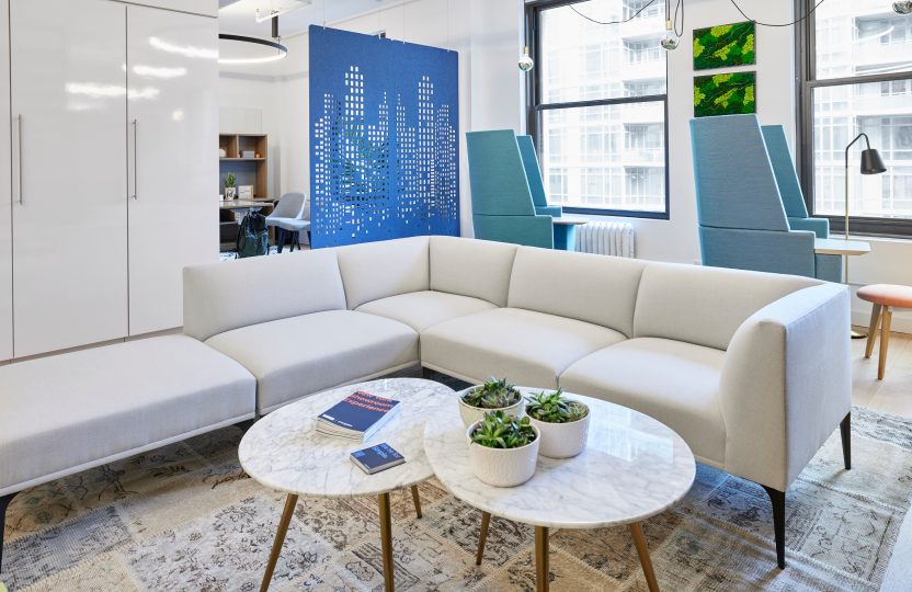 L-shaped light-gray sectional sofa, arranged with two round marble coffee tables, teal lounge chairs, and potted succulents in a modern showroom with a blue cityscape wall.